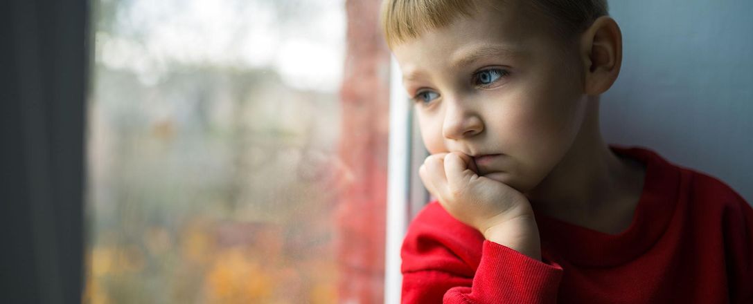 Ein kleiner Junge stützt sein Kinn auf eine Hand und schaut nachdenklich aus dem Fenster.